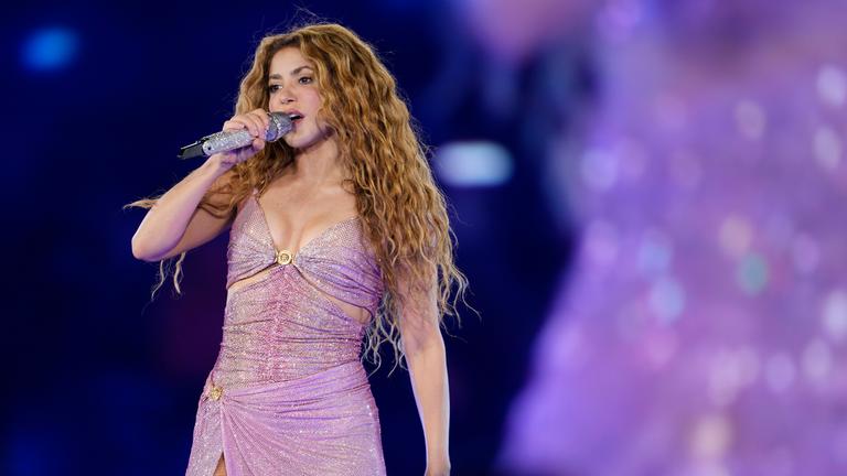 Shakira in einem lilafarbenen Outfit steht auf der Bühne und singt und performt
