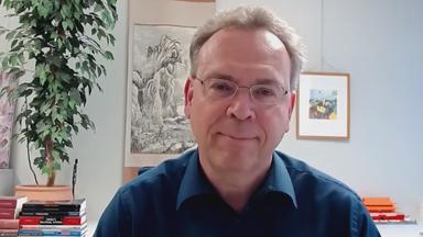 China-Experte Sebastian Heilmann von der Uni Trier per Videocall zugeschaltet. Er trägt ein schwarzes Hemd und im Hintergrund ist vermutlich ein Büroraum zu sehen, mit einer größeren Pflanze, mehreren Bildern an der Wand und Büchern auf einer Ablage zu sehen.