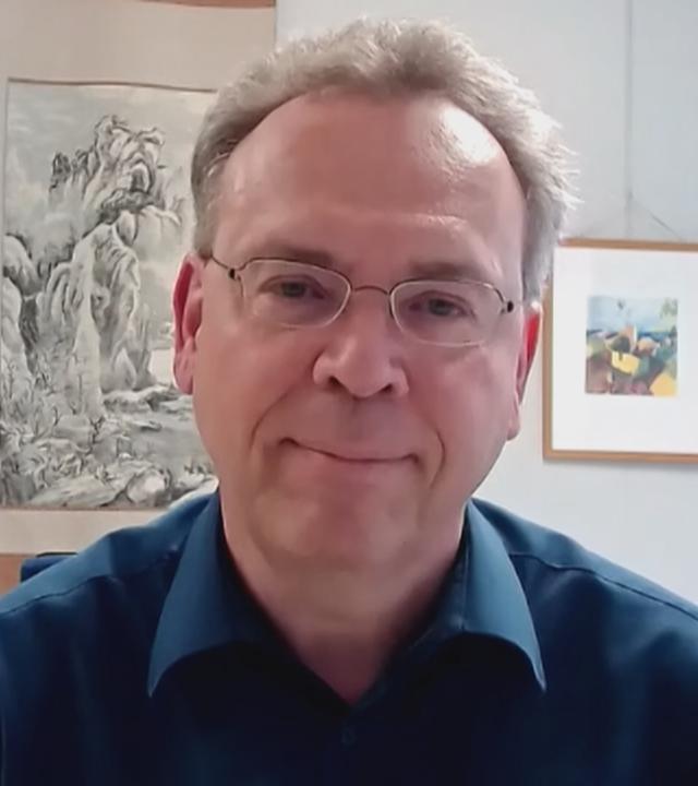 China-Experte Sebastian Heilmann von der Uni Trier per Videocall zugeschaltet. Er trägt ein schwarzes Hemd und im Hintergrund ist vermutlich ein Büroraum zu sehen, mit einer größeren Pflanze, mehreren Bildern an der Wand und Büchern auf einer Ablage zu sehen.