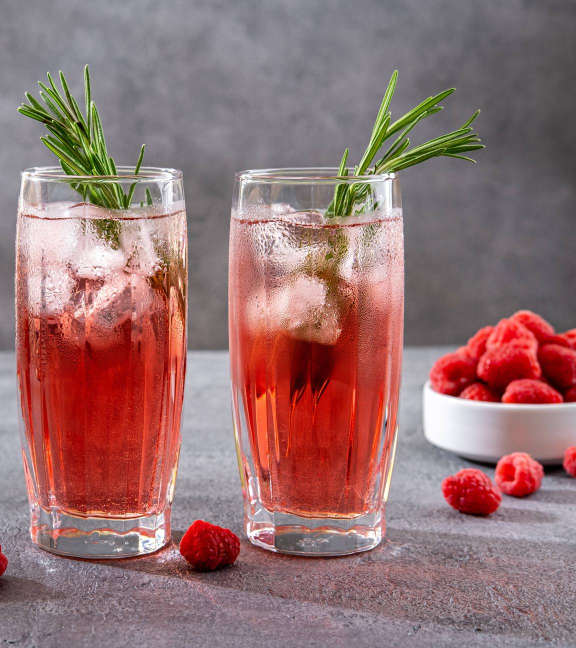 Zwei Gläser mit rötlichen Getränken, Rosmarinzweigen und Eiswürfeln stehen nebeneinander und sind umgeben von Himbeeren.