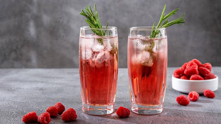 Zwei Gläser mit rötlichen Getränken, Rosmarinzweigen und Eiswürfeln stehen nebeneinander und sind umgeben von Himbeeren.