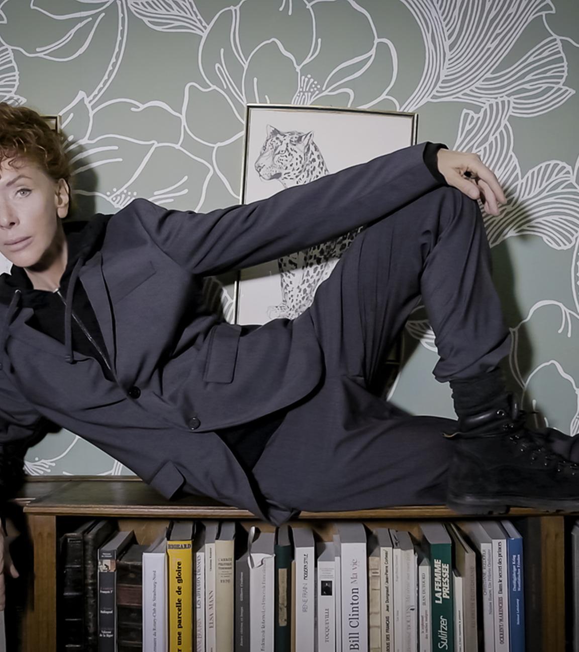 Eine Frau mit wuscheligen roten Haaren und in einem grauen Anzug liegt auf einem Bücherregal.