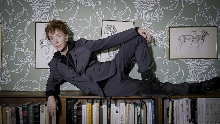 Eine Frau mit wuscheligen roten Haaren und in einem grauen Anzug liegt auf einem Bücherregal.