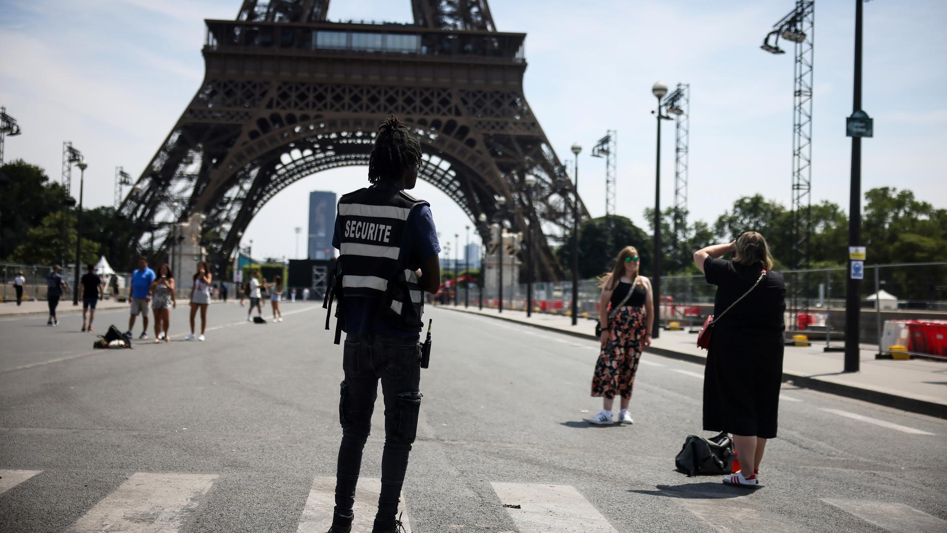20.07.2024, Frankreich, Paris: Ein Sicherheitsbeamter beobachtet Menschen, die vor dem Eiffelturm fotografiert werden.