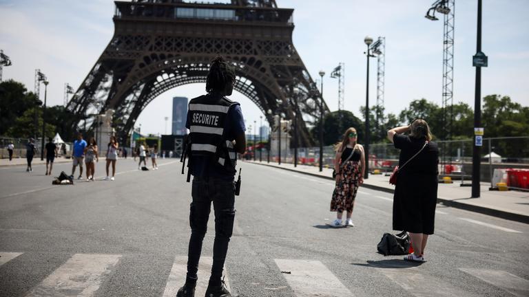 20.07.2024, Frankreich, Paris: Ein Sicherheitsbeamter beobachtet Menschen, die vor dem Eiffelturm fotografiert werden.