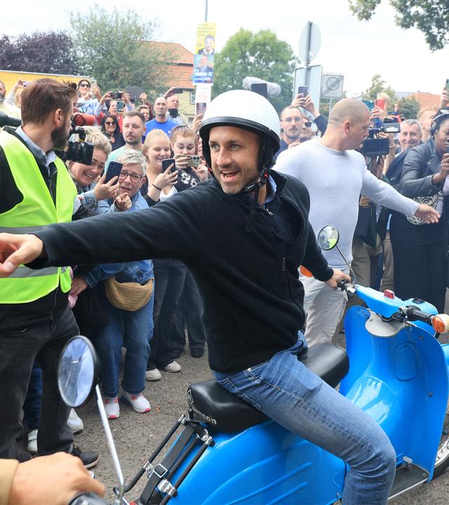 Ein Mann sitzt auf einem blauen Moped und zeigt nach links. Hiter ihm halten zwei Ordner die Menschenmenge davon ab, weiter vor zu treten. 