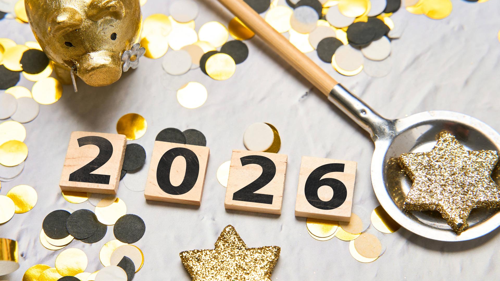 Kleine Holzplatten mit der Aufschrift "2026", Glitzersterne, Konfetti und ein Glücksschwein liegen auf einem Tisch.