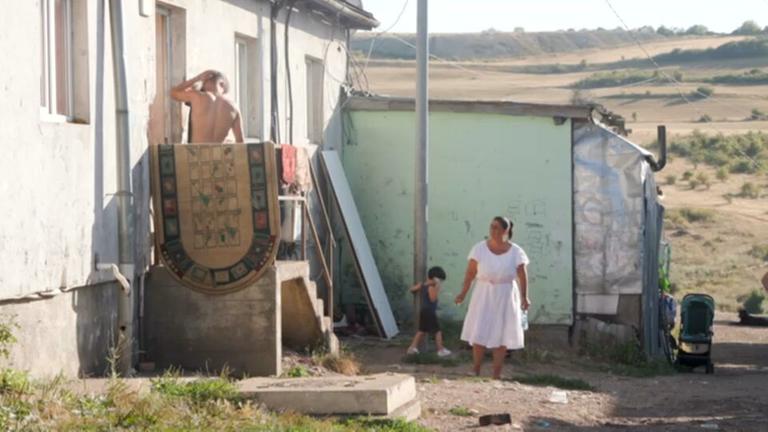 Eine Frau und ein Kind stehen vor einem Haus in der Siedlung Pata Rât in Rumänien, oben auf einer Treppe steht ein Mann mit nacktem Rücken.