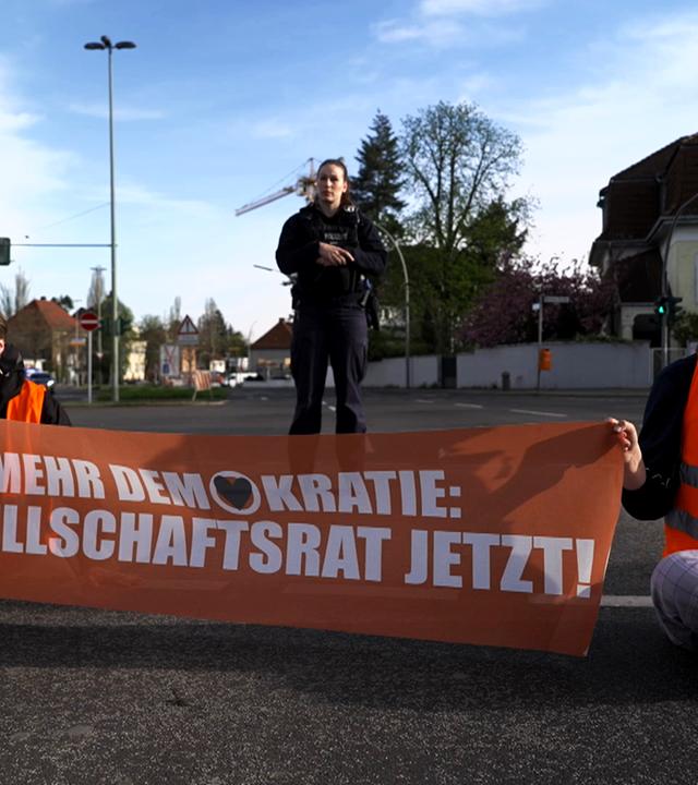 Sitzblockade Klimakleber der "Letzten Generation" an Verkehrskreuzung mit Transparent "Mehr Demokratie: Gesellschaftsrat jetzt!"
