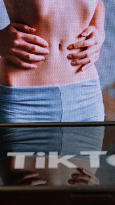 shows the logo of the Chinese social media application Tik Tok on a smartphone in front of a screen displaying an image of a skinny woman, as the "skinnytok" trend promoting thinness worries experts.