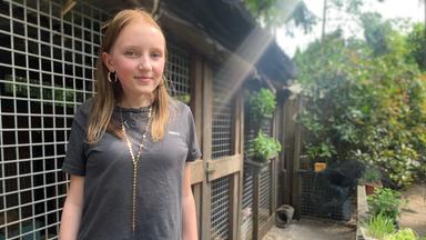 Rosa-Carmen Haasjes aus der plan b-Folge, mit durchgestrecktem Rücken in die Kamera lächelnd, steht im Garten vor einem Schuppen.