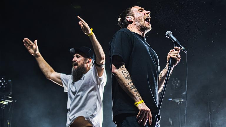 Links im Bild steht ein Mann, mit dunklem Bart und schwarzer Kappe auf dem Kopf. Er trägt ein graues T-Shirt und hebt seine beiden Arme in die Luft. Sein Blick geht nach links. Rechts daneben steht ein weiterer Mann, dessen Blick in die andere Richtung geht und in ein Mikrofon singt. Er trägt ein schwarzes T-Shirt und auf seinem Arm sind Tattoos zu erkennen. Beide stehen auf einer Bühne.