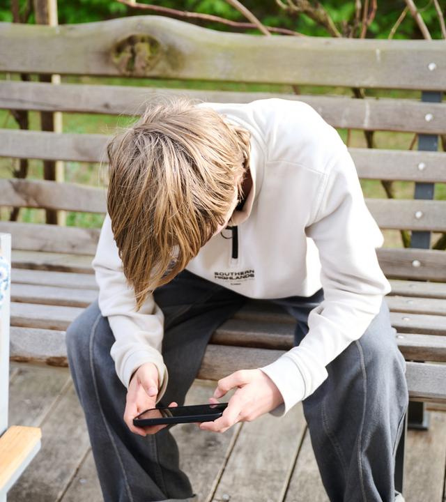 Bayern, Roth: Ein Junge guckt auf sein Smartphone, während er auf einer Bank in einem Garten sitzt. Symbolbild