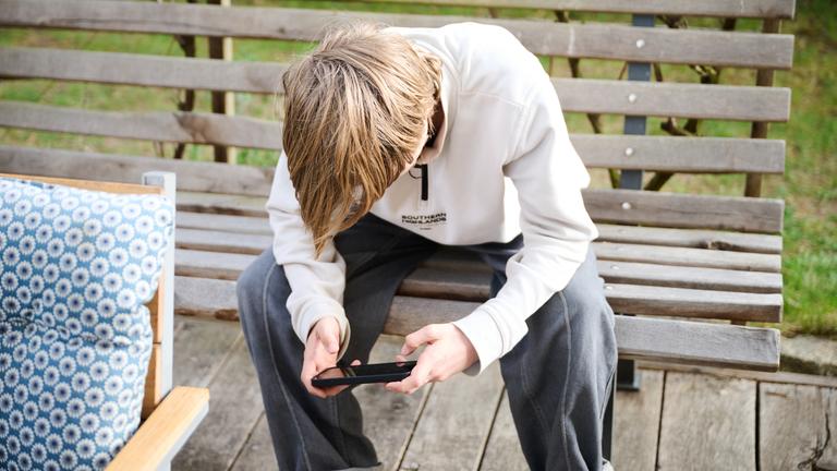 Bayern, Roth: Ein Junge guckt auf sein Smartphone, während er auf einer Bank in einem Garten sitzt. Symbolbild