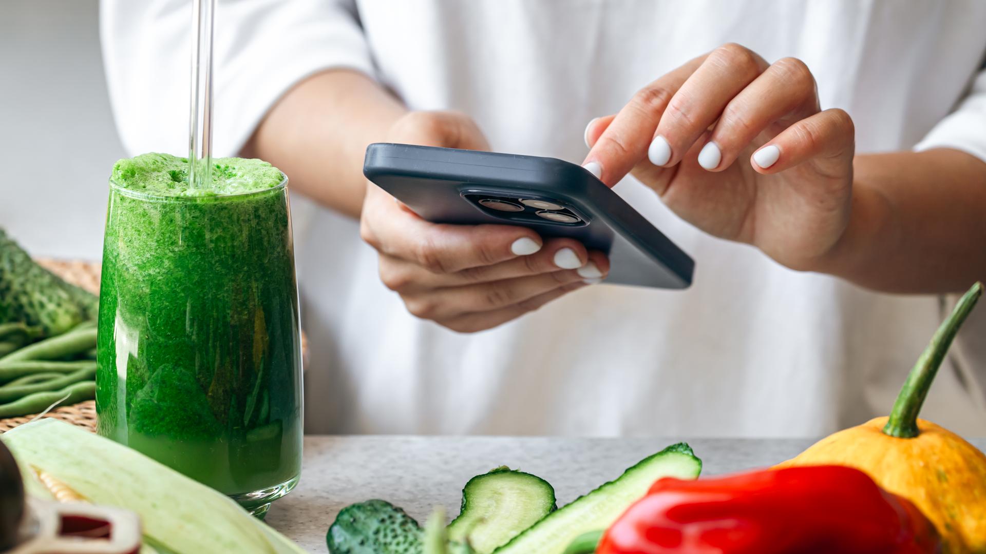 Ein grüner Smoothie steht auf einem Tisch, auf dem Gemüse liegt. Dahinter steht eine eine Frau, die auf einem Smartphone tippt. 