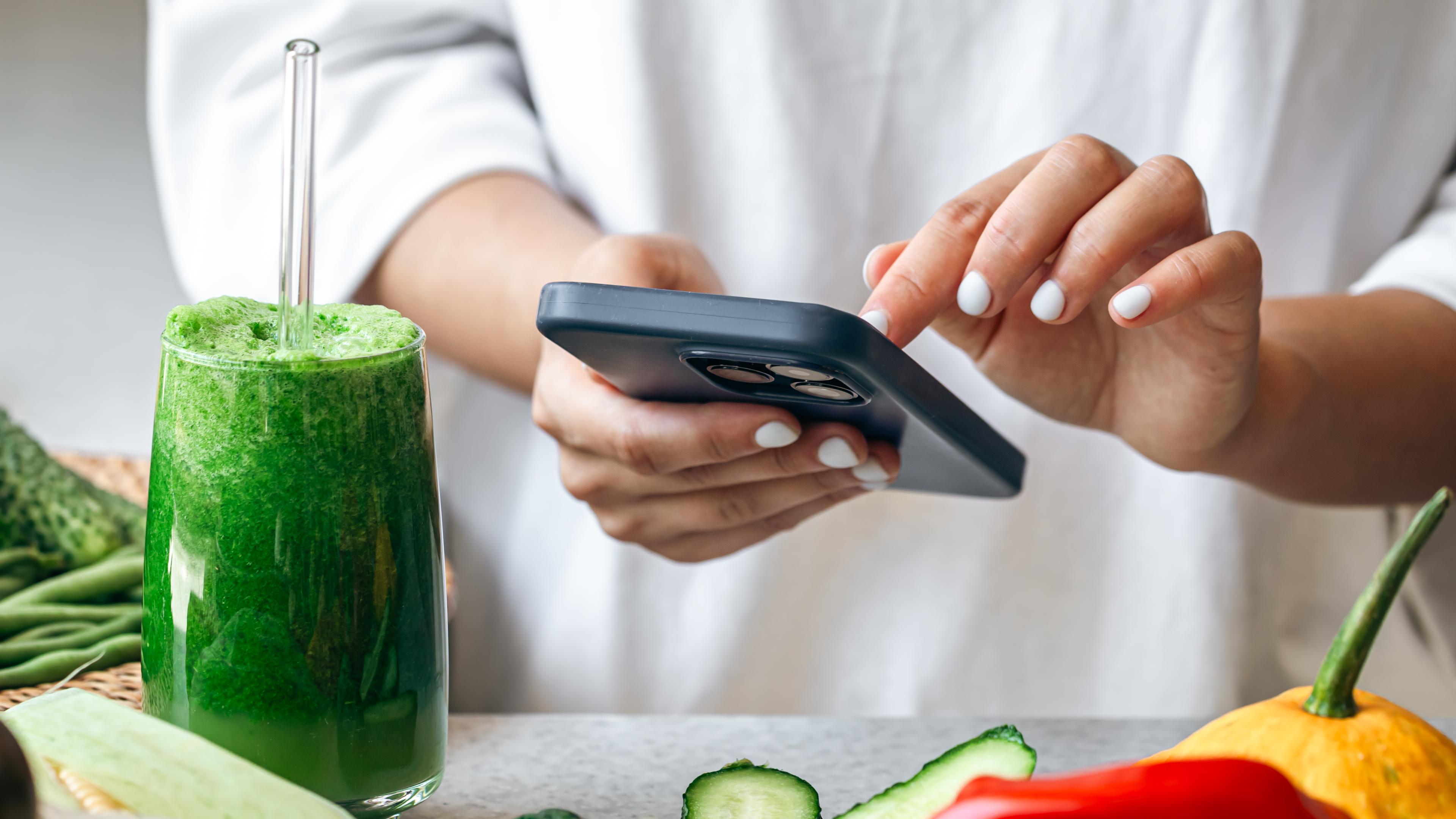Ein grüner Smoothie steht auf einem Tisch, auf dem Gemüse liegt. Dahinter steht eine eine Frau, die auf einem Smartphone tippt. 