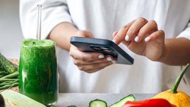 Ein grüner Smoothie steht auf einem Tisch, auf dem Gemüse liegt. Dahinter steht eine eine Frau, die auf einem Smartphone tippt. 