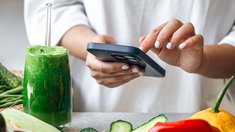 Ein grüner Smoothie steht auf einem Tisch, auf dem Gemüse liegt. Dahinter steht eine eine Frau, die auf einem Smartphone tippt. 