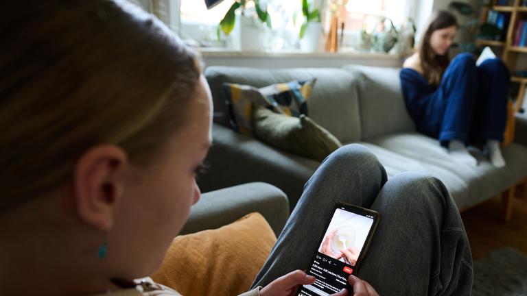 Ein Mädchen nutzt ihr Smartphone, während ein anderes Mädchen ein Buch liest.