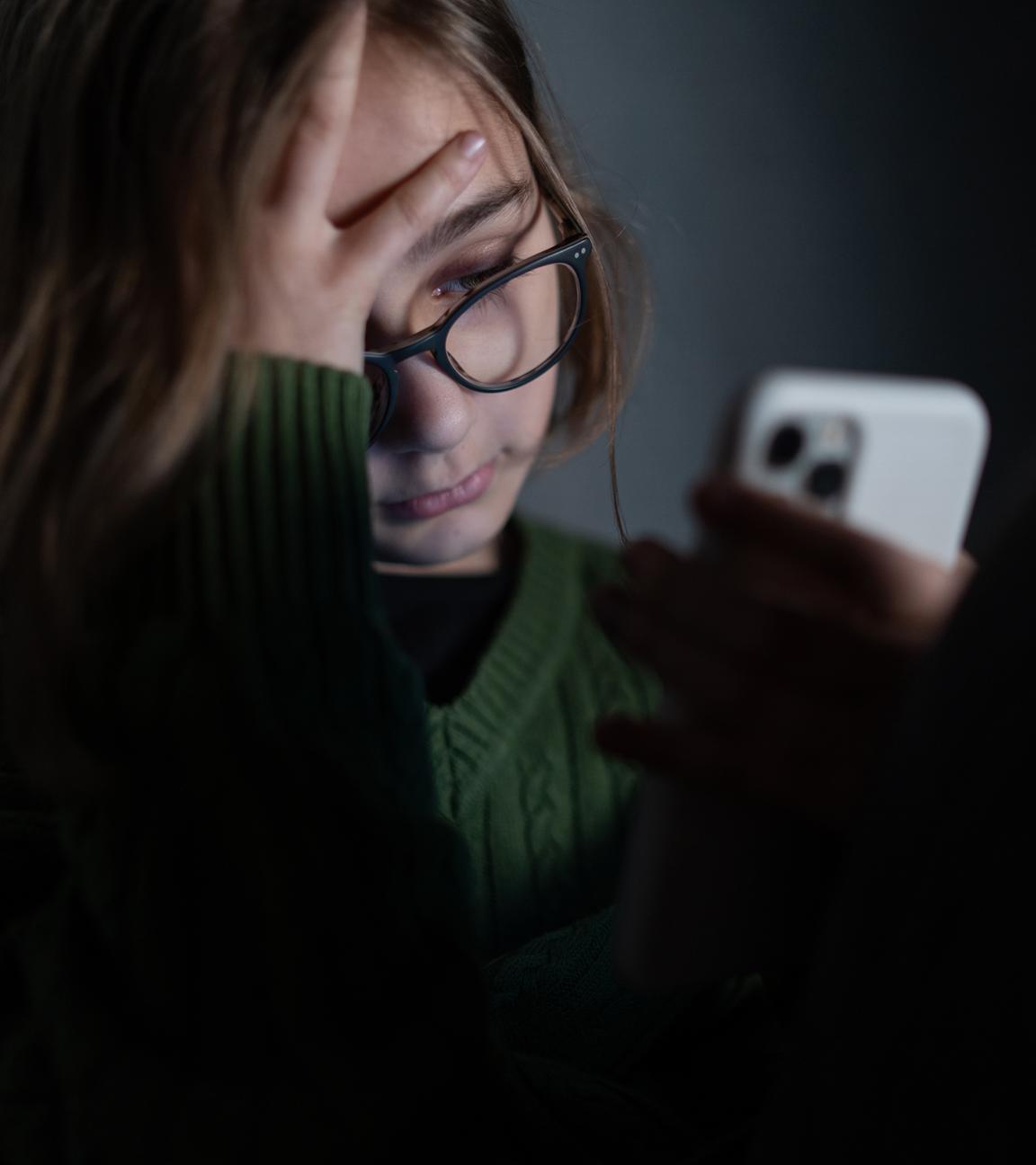 Ein Mädchen sitzt im Dunkeln und stützt ihr Gesicht auf ihre Hand. Dabei schaut sie traurig auf ihr Handy-Display.