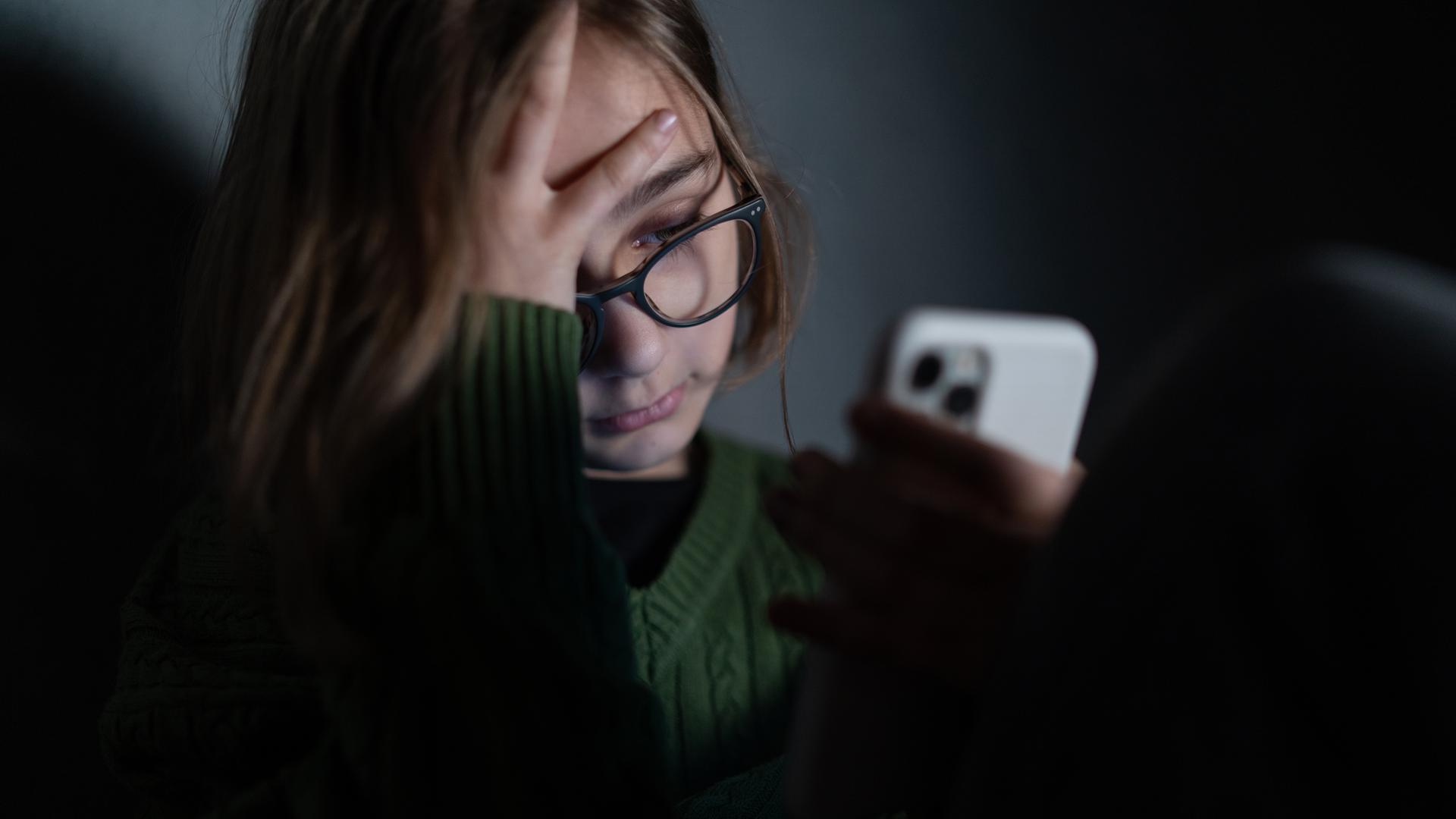 Ein Mädchen sitzt im Dunkeln und stützt ihr Gesicht auf ihre Hand. Dabei schaut sie traurig auf ihr Handy-Display.