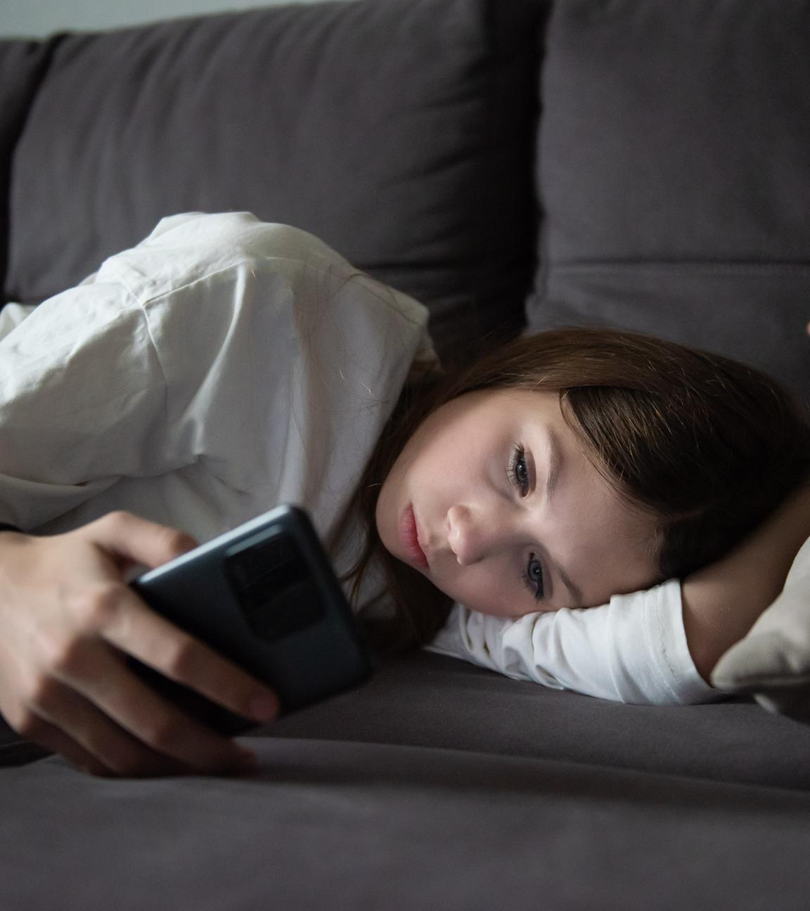 Ein trauriges Teenager-Mädchen liegt mit einem Smartphone in der Dunkelheit. 