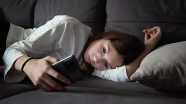 Ein trauriges Teenager-Mädchen liegt mit einem Smartphone in der Dunkelheit. 