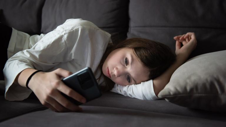 Ein trauriges Teenager-Mädchen liegt mit einem Smartphone in der Dunkelheit. 