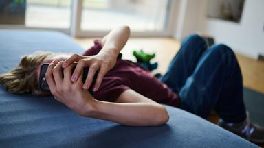 Ein Teenager guckt auf ein Handy, während er auf Boden auf einer Matratze liegt. 