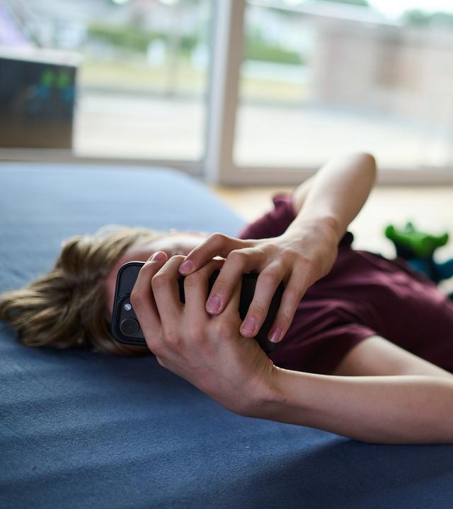 Ein Teenager guckt auf ein Handy, während er auf Boden auf einer Matratze liegt. 