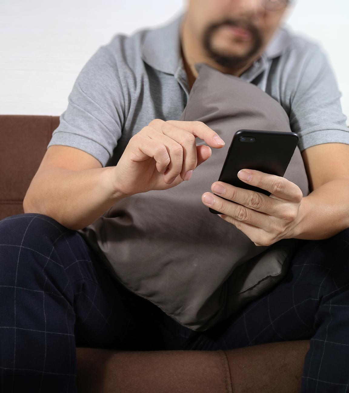 ein Mann sitzt auf dem Sofa und tippt auf seinem Handy. Sein Kopf ist nicht zu sehen.
