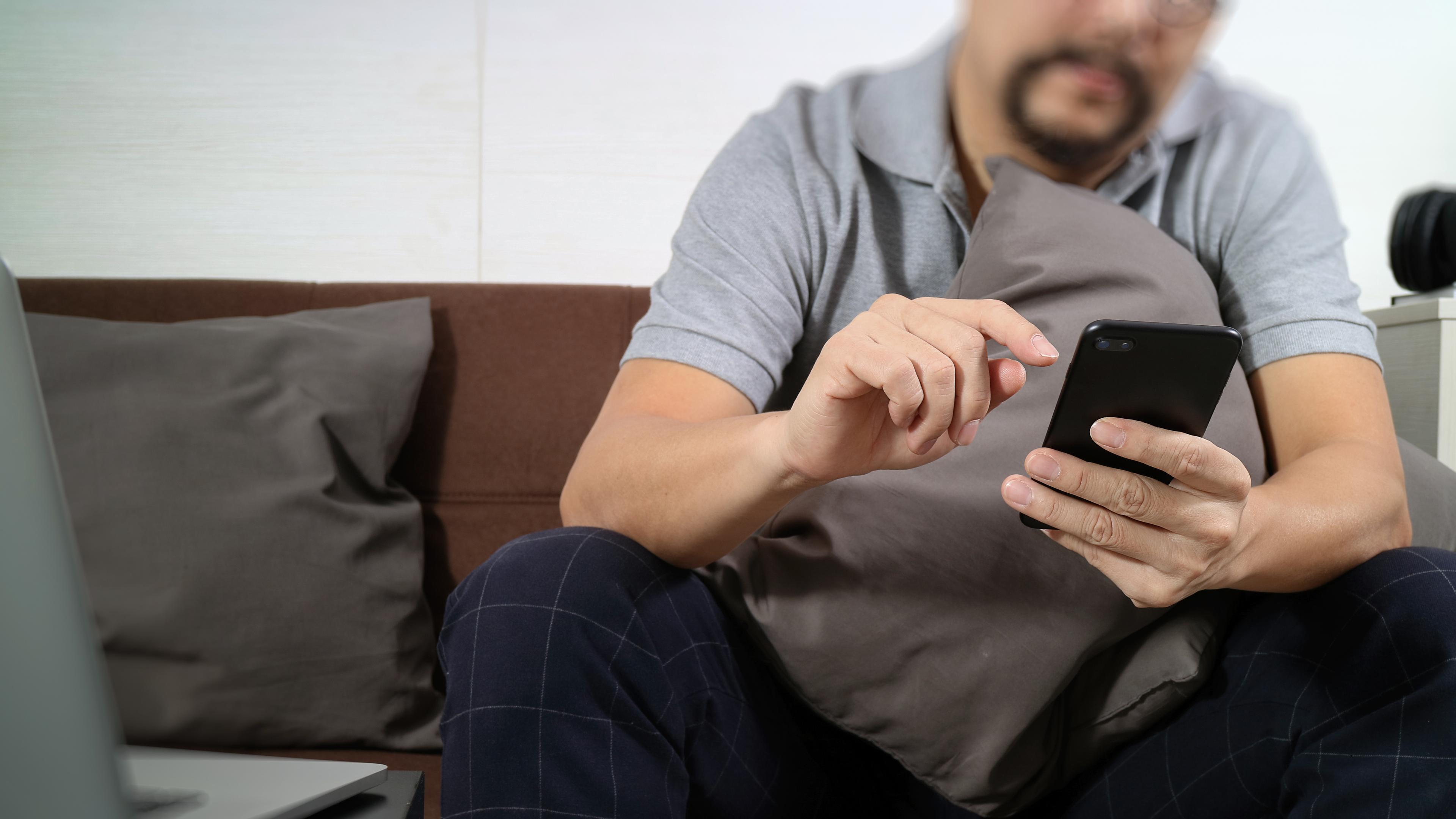 ein Mann sitzt auf dem Sofa und tippt auf seinem Handy. Sein Kopf ist nicht zu sehen.