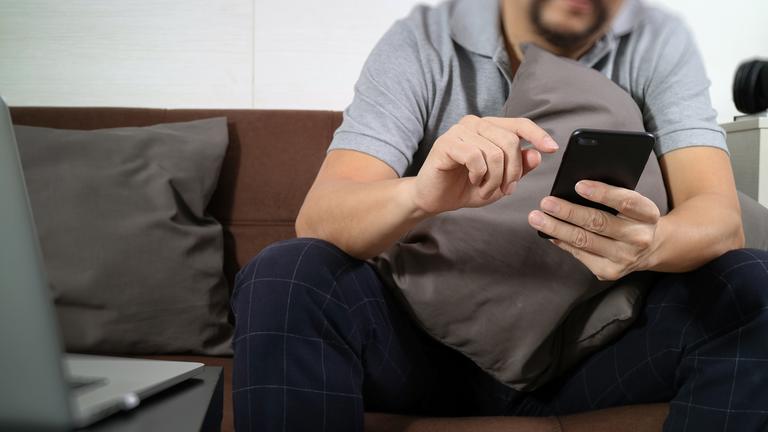 ein Mann sitzt auf dem Sofa und tippt auf seinem Handy. Sein Kopf ist nicht zu sehen.