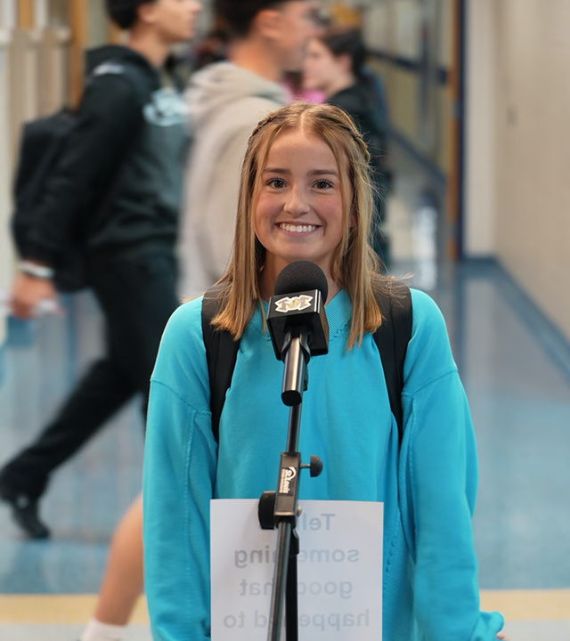 Eine Schülerin steht auf einem Schulgang vor einem Mikrofon und lächelt.