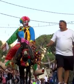 Ein Esel wird von einem Mann geritten, der als Clown verkleidet ist. Der Esel ist ebenfalls bunt geschmückt.