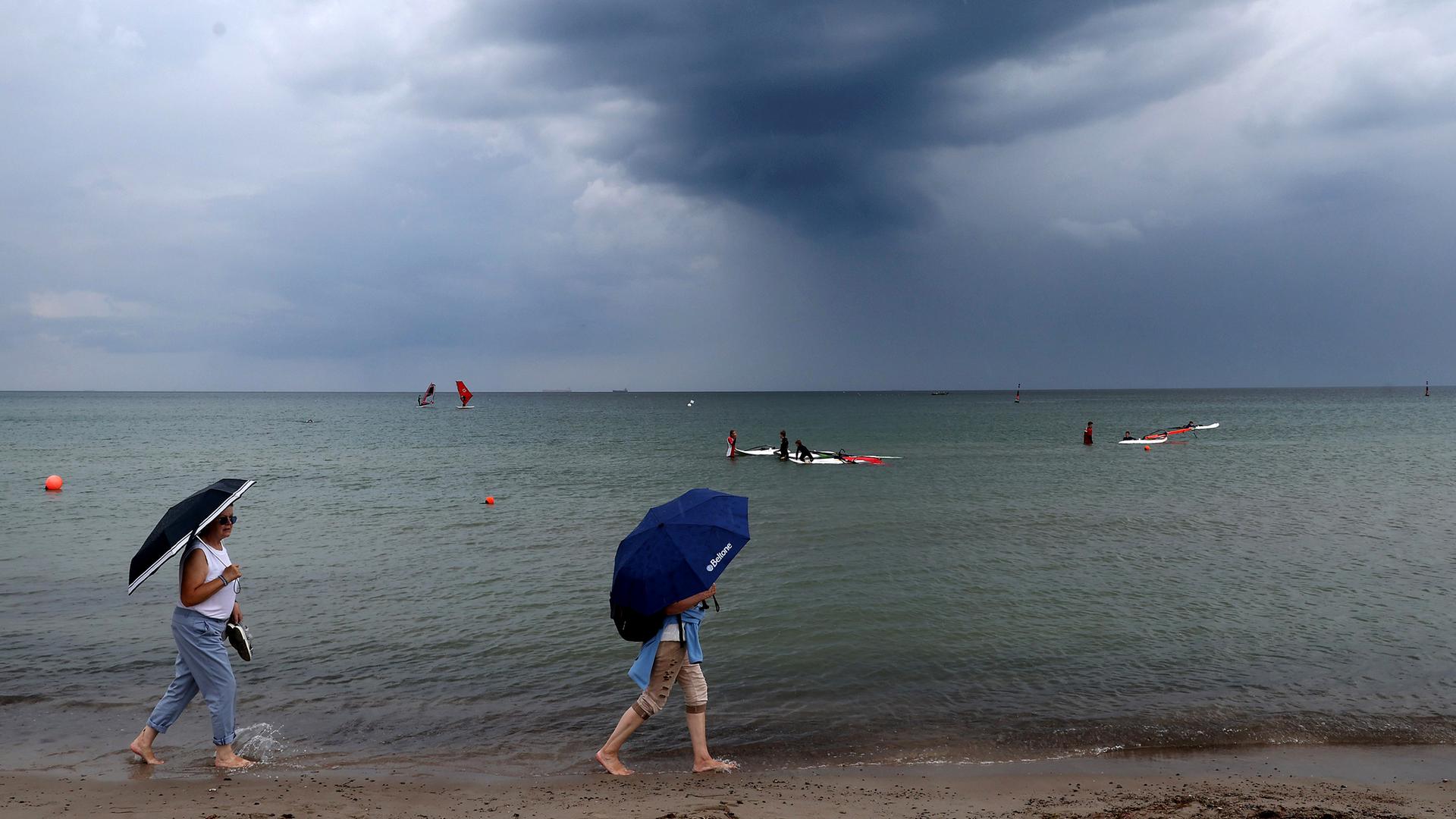 Über der Ostsee ziehen dunkle Regenwolken auf, während zwei Frauen mit Regenschirmen am Strand unterwegs sind