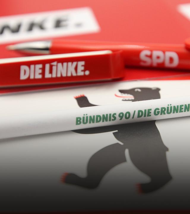 Symbolbild: Auf einer Flagge von Berlin liegen Kugelschreiber der Parteien die Linke, SPD und Bündnis 90 Die Grünen. 