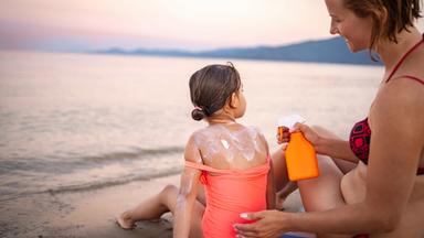 Mutter cremt Tochter am Strand mit Sonnencreme ein.