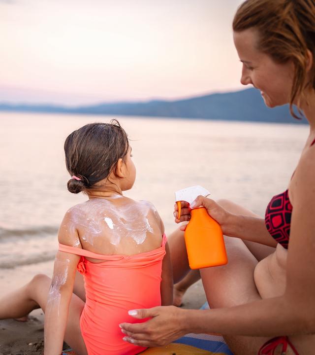 Mutter cremt Tochter am Strand mit Sonnencreme ein.
