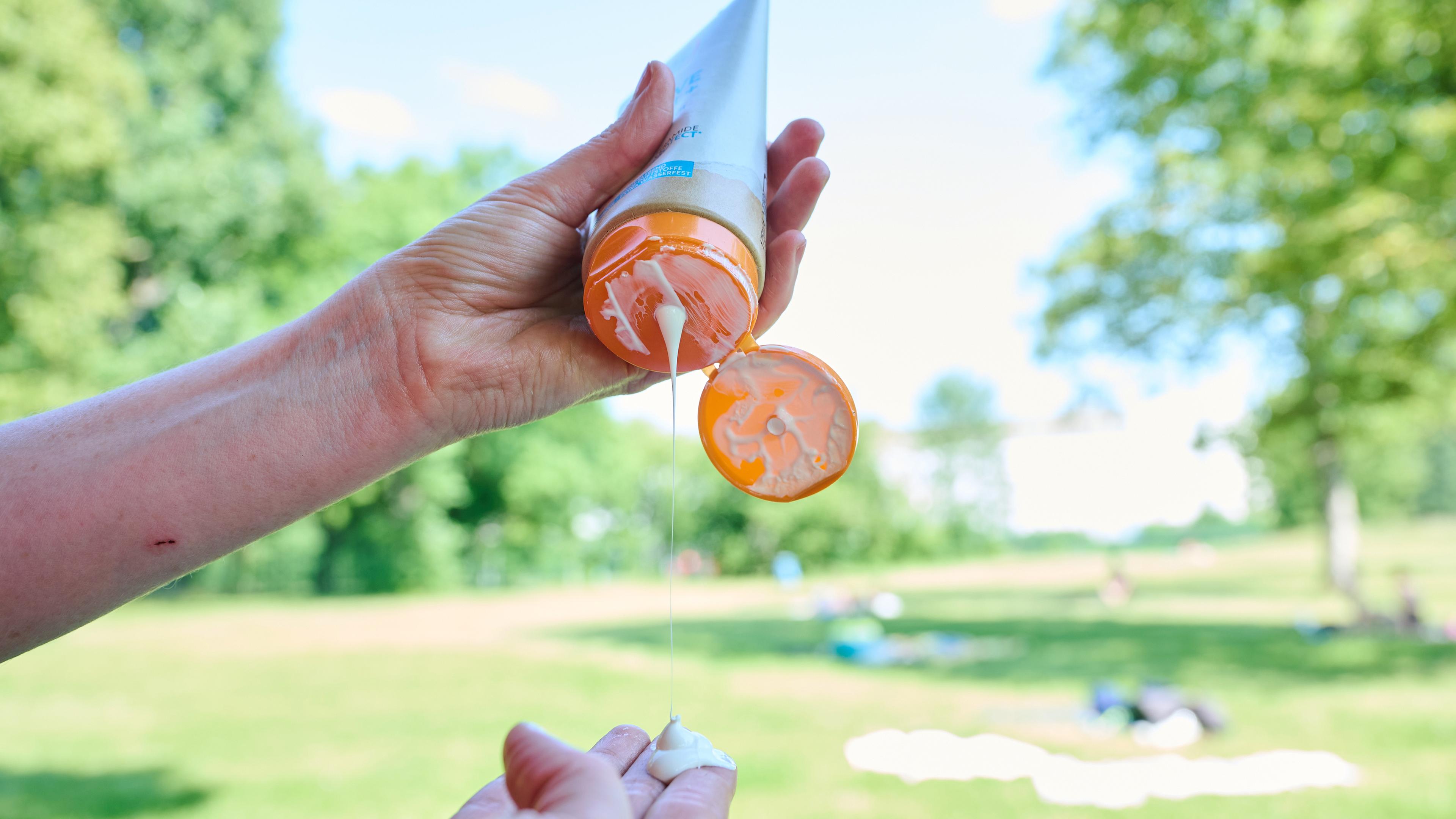 Auf dem Bild sind die Hände einer Person zu sehen, in der linken Hand hält sie eine Sonnencreme. Die Sonnenmilch ist geöffnet und es tropft Creme auf die ausgestreckten Finger der anderen Hand. Im Hintergrund ist ein Park bei sonnigem Wetter zu sehen.