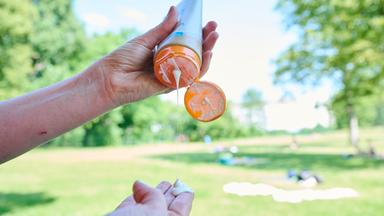 Auf dem Bild sind die Hände einer Person zu sehen, in der linken Hand hält sie eine Sonnencreme. Die Sonnenmilch ist geöffnet und es tropft Creme auf die ausgestreckten Finger der anderen Hand. Im Hintergrund ist ein Park bei sonnigem Wetter zu sehen.