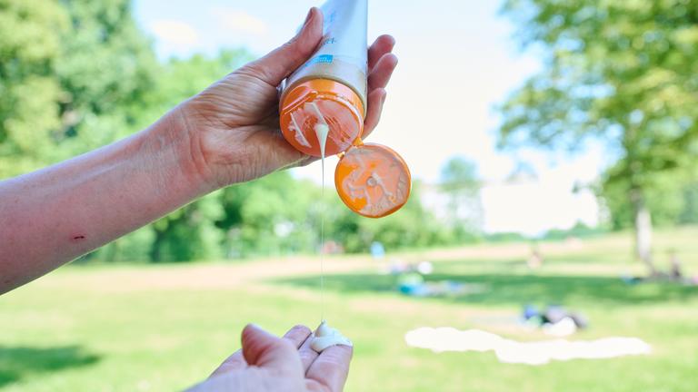 Auf dem Bild sind die Hände einer Person zu sehen, in der linken Hand hält sie eine Sonnencreme. Die Sonnenmilch ist geöffnet und es tropft Creme auf die ausgestreckten Finger der anderen Hand. Im Hintergrund ist ein Park bei sonnigem Wetter zu sehen.