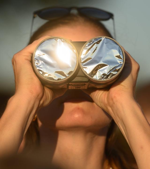 Eine Frau schaut mit einem Schutzfilter über dem Fernglas in richtung der partiellen Sonnenfinsternis am 12.08.2026.