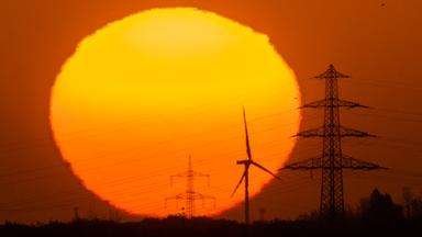 Eine große rote Sonne geht hinter einer Landschaft mit Strommasten und Windrädern unter. 