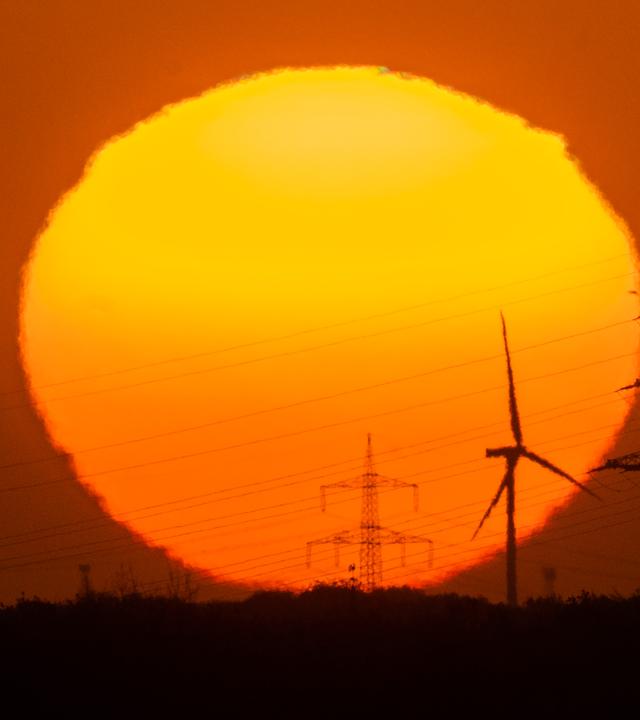 Eine große rote Sonne geht hinter einer Landschaft mit Strommasten und Windrädern unter. 