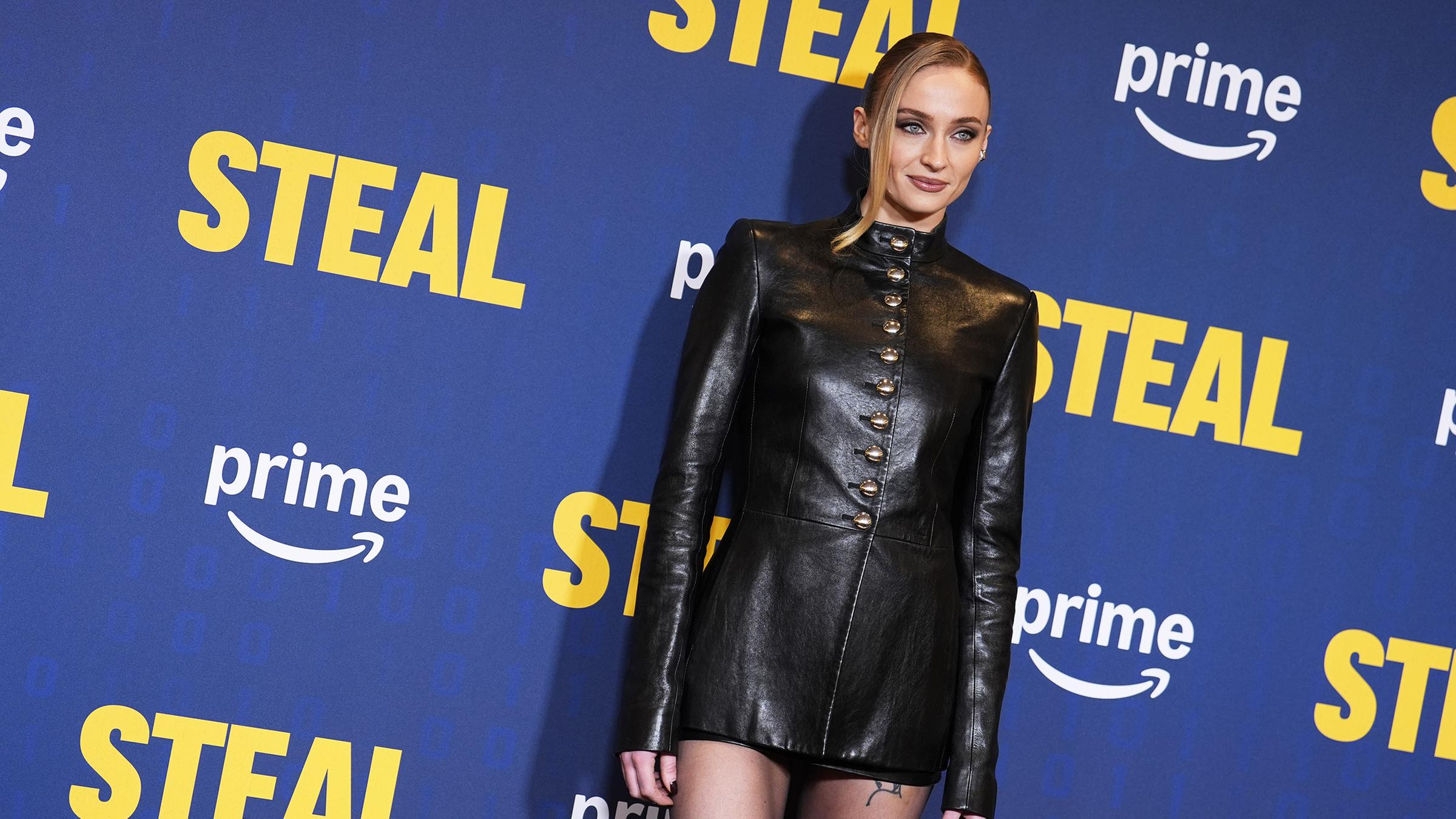 Sophie Turner steht vor einer Fotowand und posiert. Sie trägt ein schwarzes Kleid.