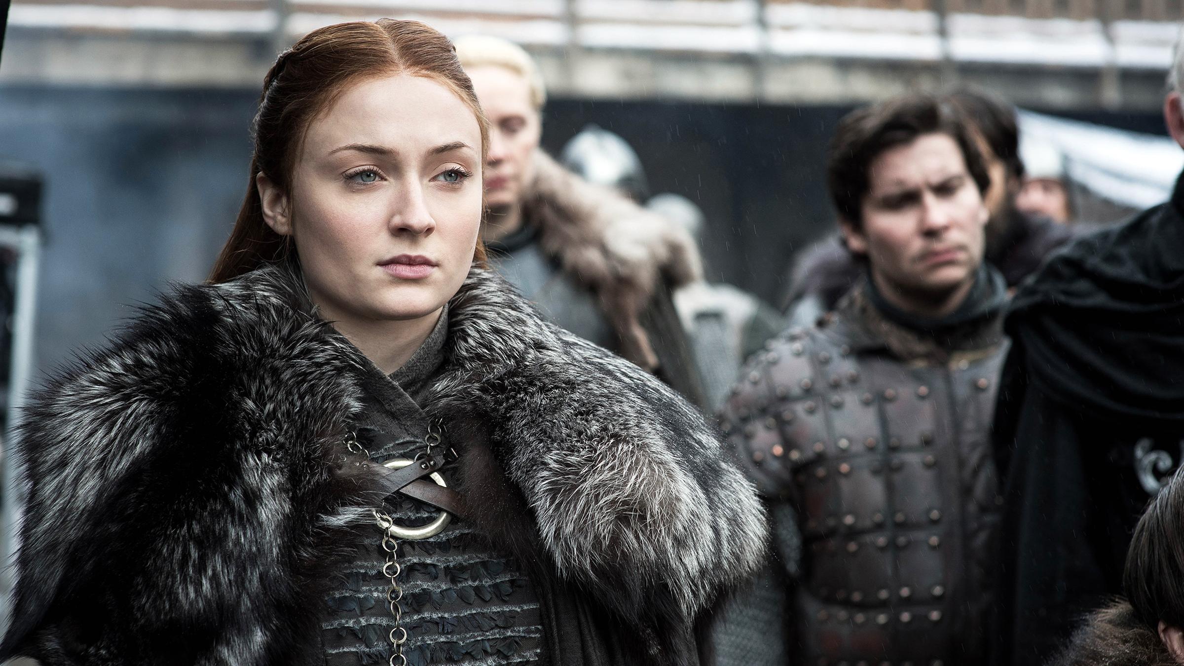 Sophie Turner als “Sansa Stark“ in der Serie "Game of Thrones" 2019. Sie hat einen Pelz umhängen und ist von Menschen umgeben.