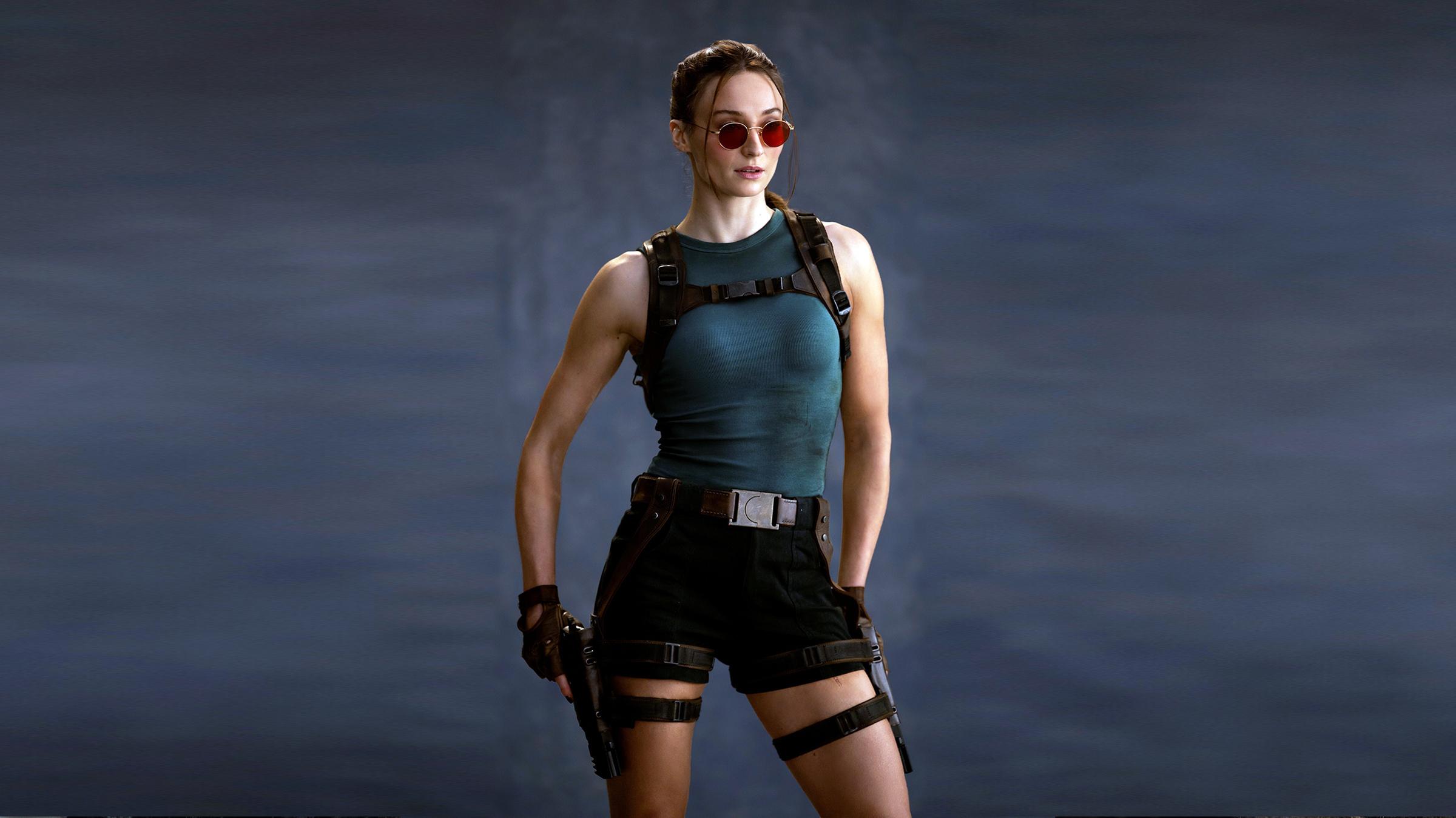 Sophie Turner in ihrer Rolle als “Lara Croft“. Sie trägt eine Sonnenbrille und hat eine Waffe in der Hand.
