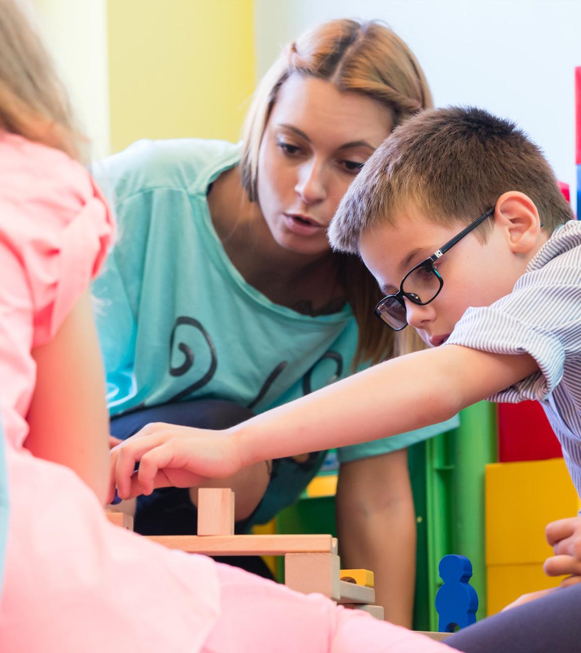 Vorschuljunge arbeitet gemeinsam mit seinen Spielkameraden am Bau einer Struktur aus Holzbausteinen – unter der Anleitung einer jungen Kindergärtnerin.