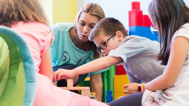 Vorschuljunge arbeitet gemeinsam mit seinen Spielkameraden am Bau einer Struktur aus Holzbausteinen – unter der Anleitung einer jungen Kindergärtnerin.